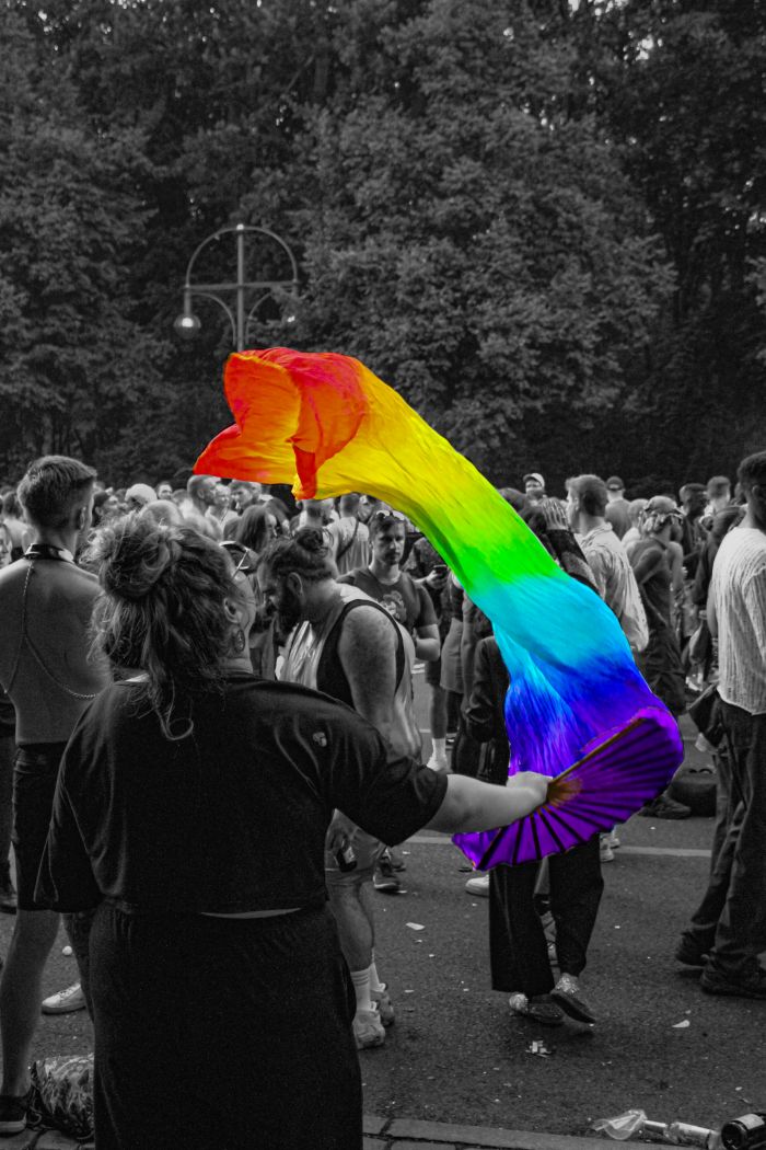 Eine Person auf dem CSD mit einem Fächer. Ein am fächer befestigtes Buntes Band fliegt durch die Luft. Der Rest ist in Graustufen